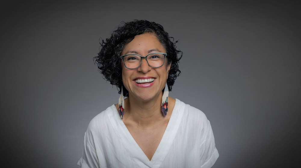 Portrait of a smiling woman with glasses and curly hair wearing a white top and earrings