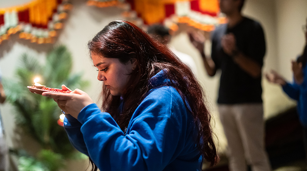 Person holding a lit diya during a Hindu prayer ritual, with others in the background

