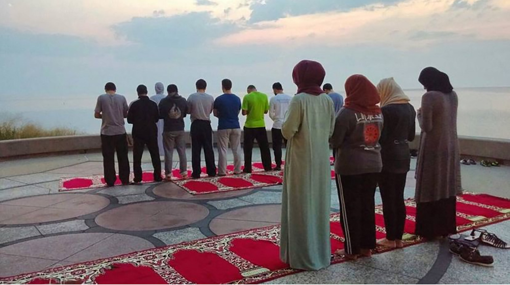 Group of people standing on prayer rugs outdoors, participating in a Muslim prayer facing the water
