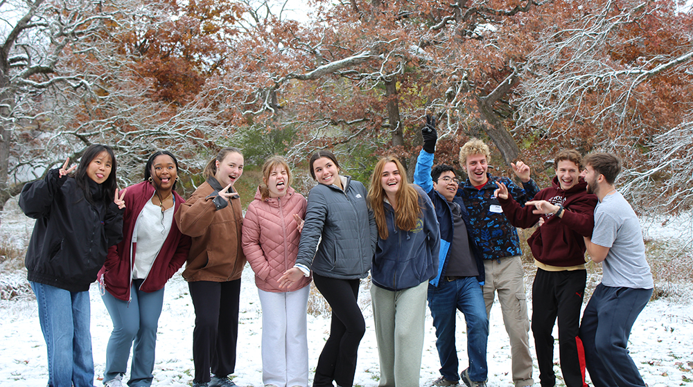 Group of students from Campus Ministry standing together outdoors in the snow, smiling and posing playfully with autumn-colored trees in the backgroun