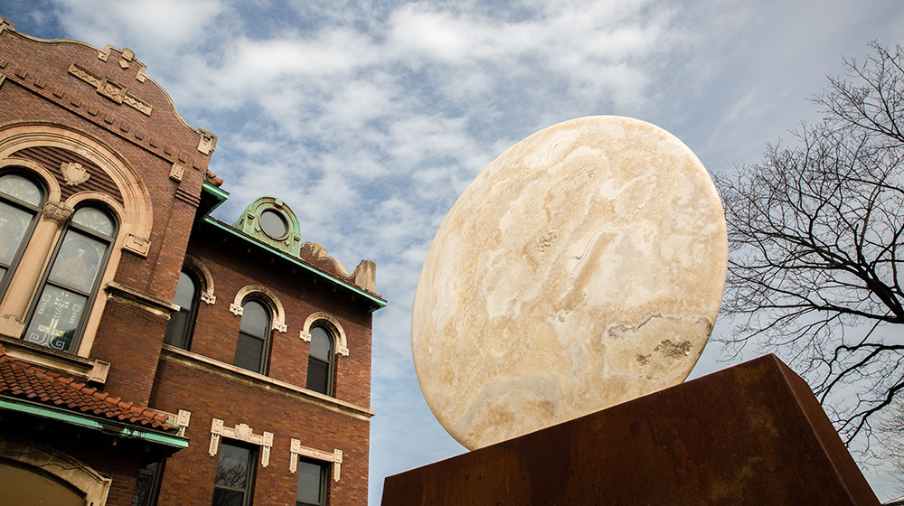 Large circular stone sculpture beside a historic brick building under a cloudy sky