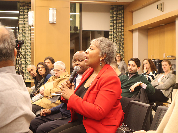 Woman in a red blazer speaking into a microphone during a group discussion with seated audience