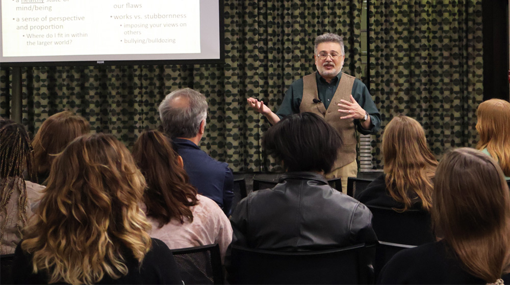 Speaker presenting to a seated audience with a slide projected behind him