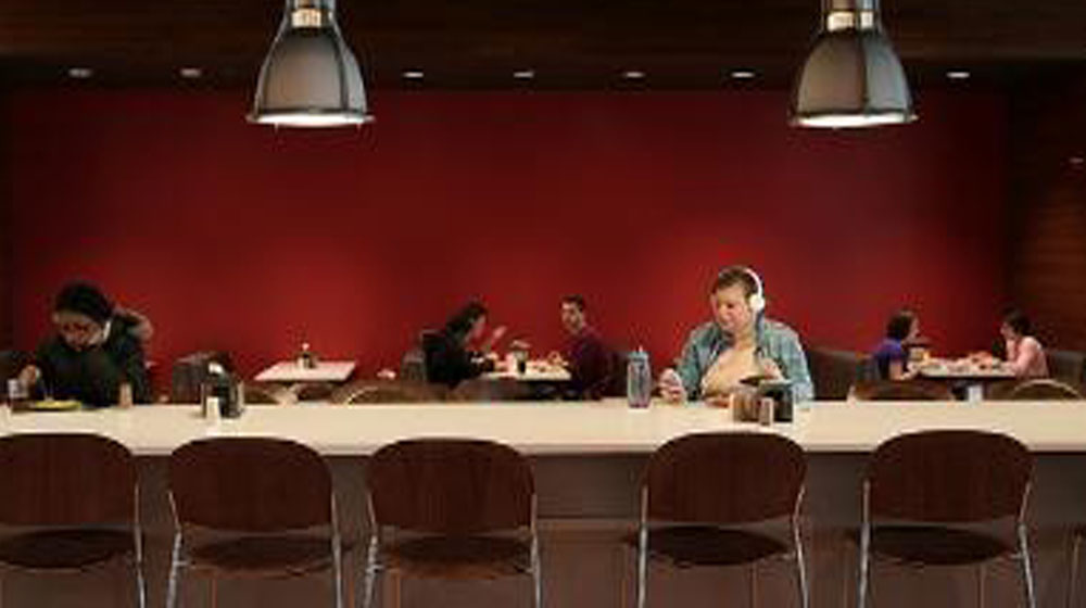 Students seated at tables and a long counter in a dining area with a red wall, some eating and studying.