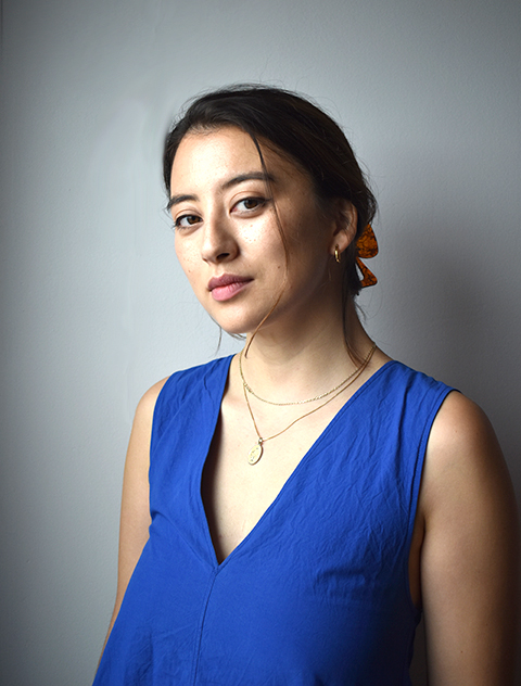 Woman in a blue top with hair tied back, posing against a plain background.