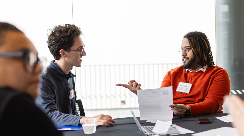 Two faculty or staff member wearing name tags and seated at a table, discuss topics on a handout during a conference session.