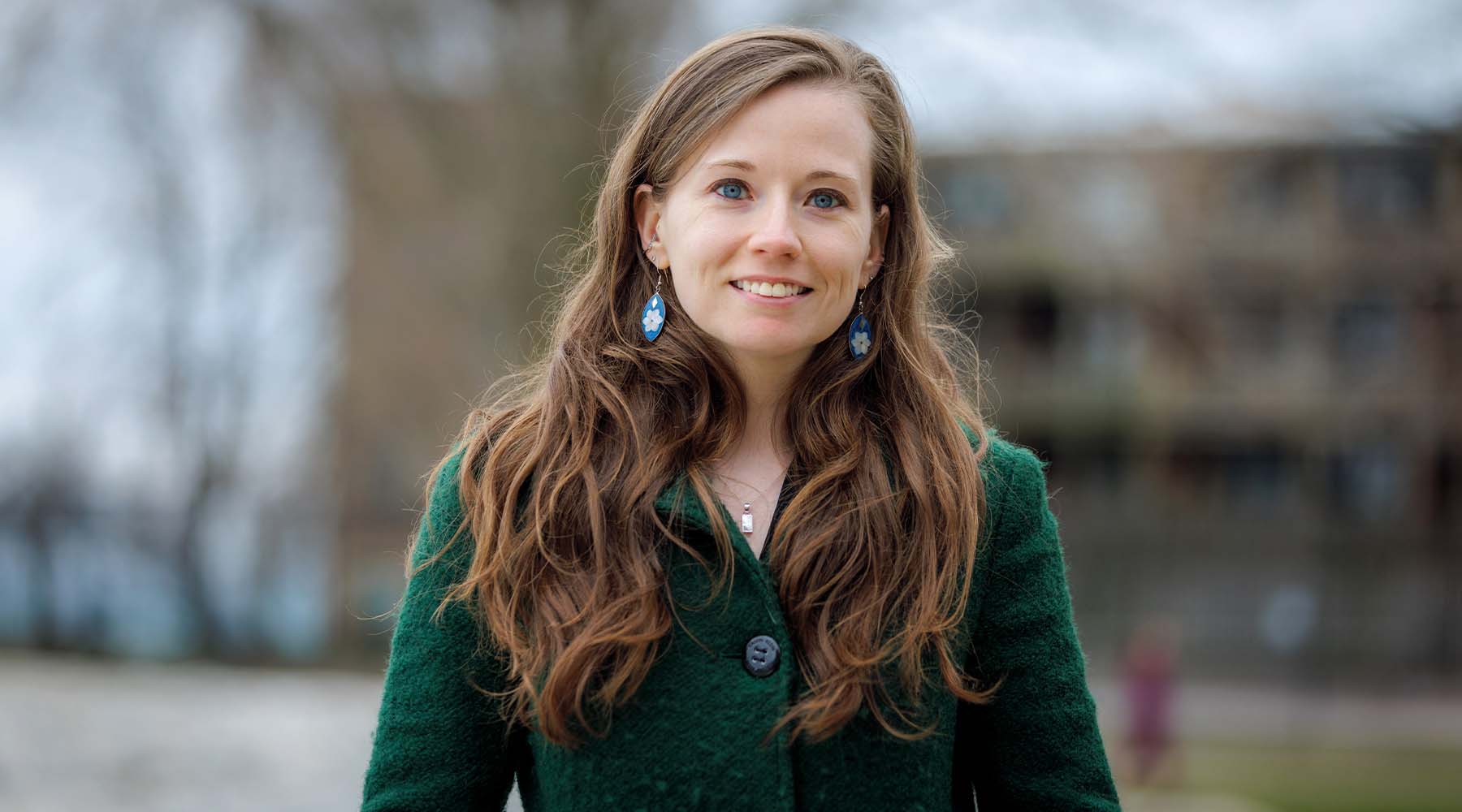 Portrait of a woman with long brown hair wearing a green coat, standing outdoors with a softly blurred campus background.