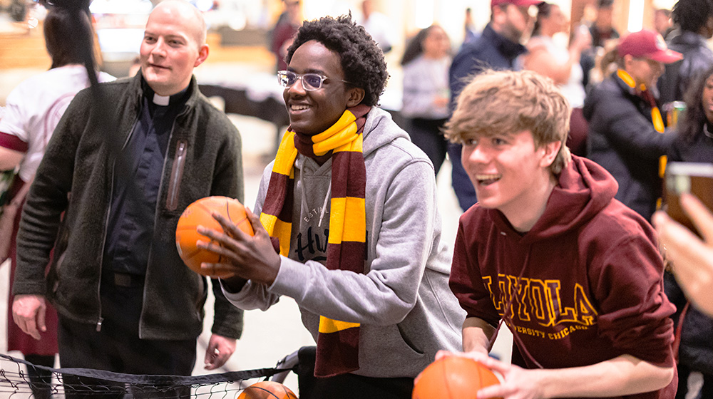 Two students playing a basketball shooting game with a Jesuit standing in the background