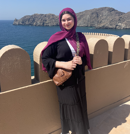 Woman wearing a purple headscarf standing by a seaside overlook with rocky cliffs in the background.
