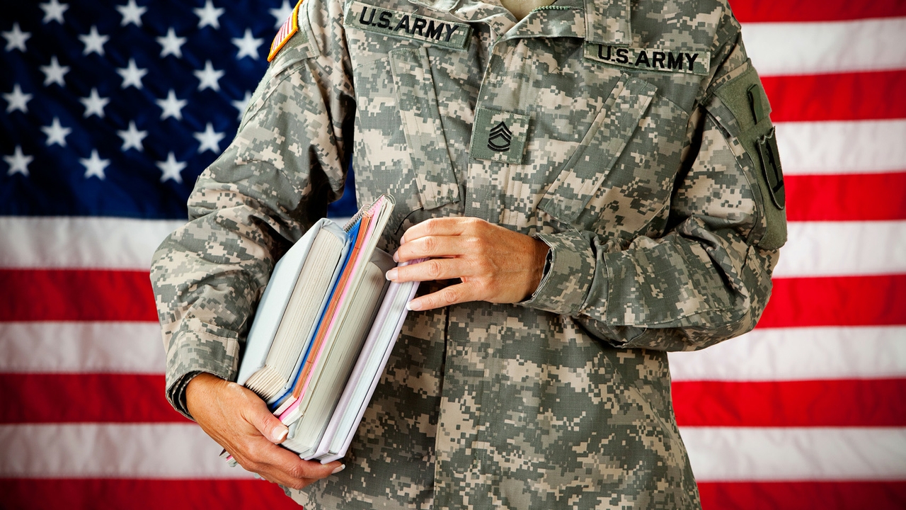 US military personnel in uniform holding books for school. The person is standing in front of an American flag backdrop.