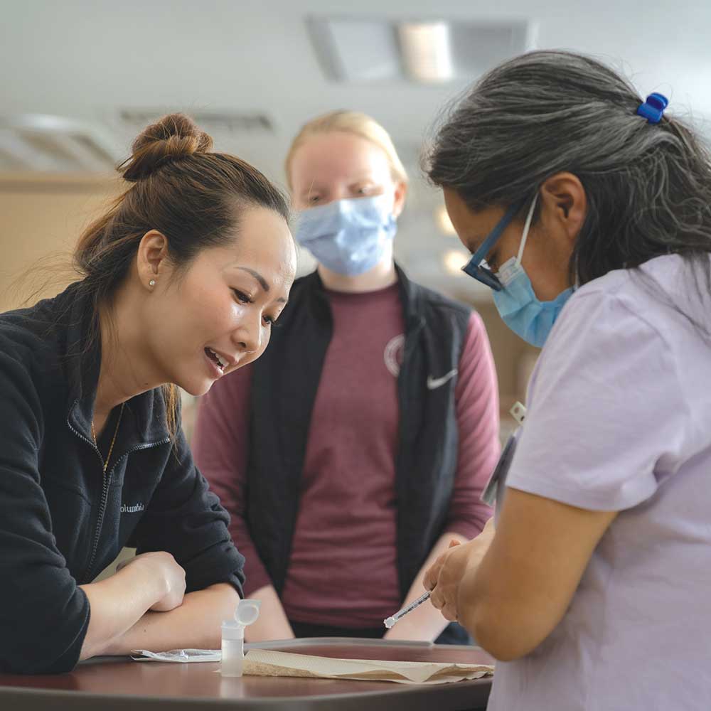 Two individuals observe as another prepares a syringe at a table, with medical supplies and paperwork visible in a clinical setting.