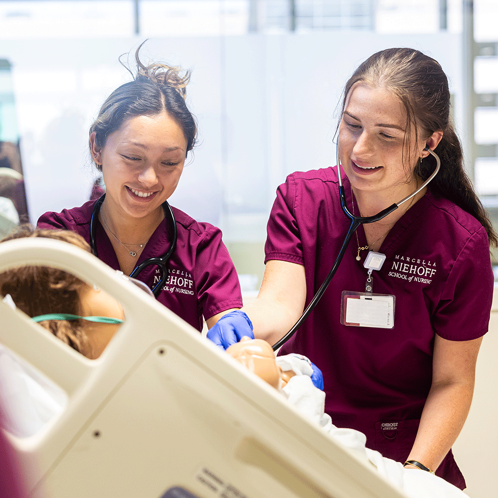 Two female students in maroon scrubs smile and look down at a manikin