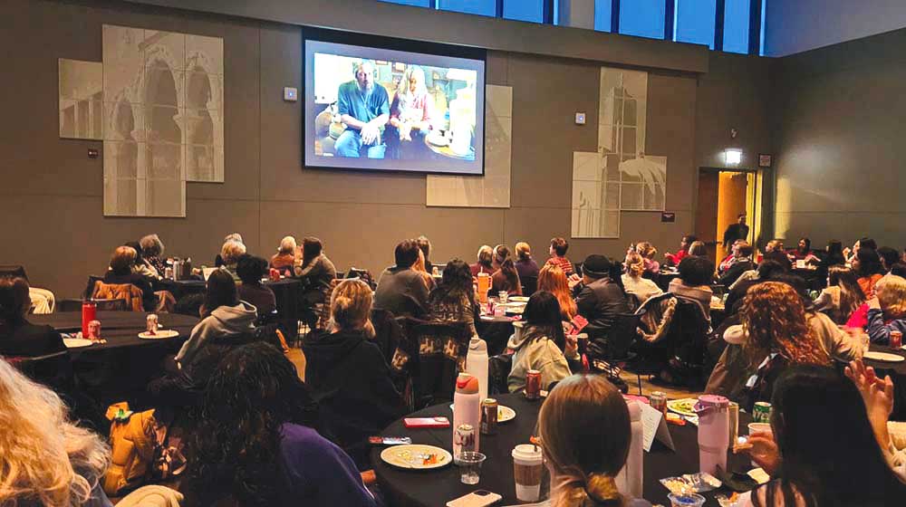 Audience seated at round tables watches a projected video during an indoor event, with plates, drinks, and notebooks visible.