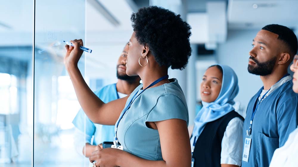 Adults in blue scrubs look on as a woman writes on a board