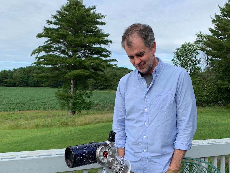 Man standing outdoors by a telescope on a deck, looking down, with trees and a grassy field in the background.