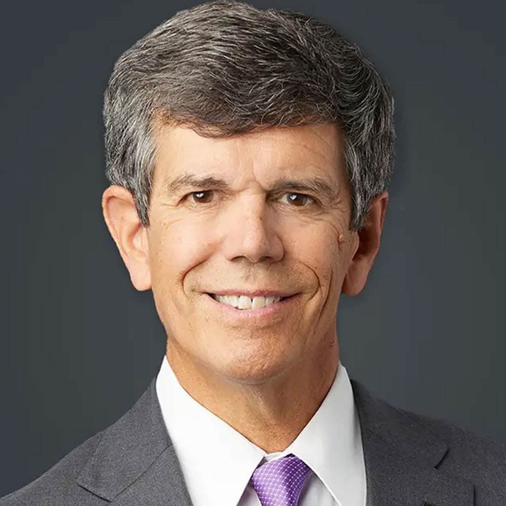 A man smiles for a portrait against a grey background. He is wearing a grey suit, white shirt, and a lavender tie.