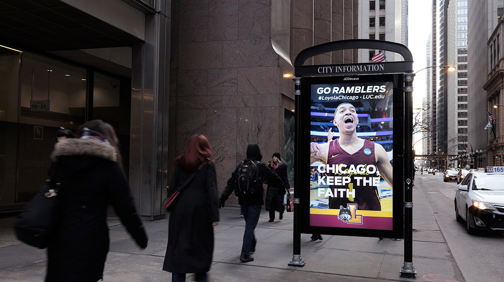 Pedestrians walk along a downtown sidewalk past a digital kiosk displaying a Loyola Chicago sports advertisement