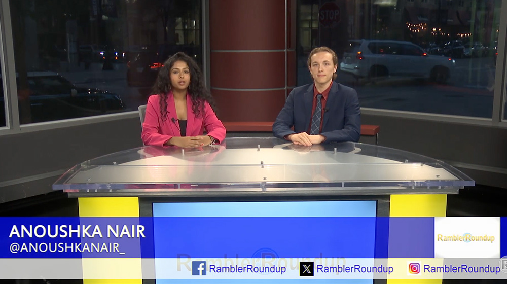 Two news anchors sit at a studio desk delivering a broadcast, with graphics displaying their names and social media information.
