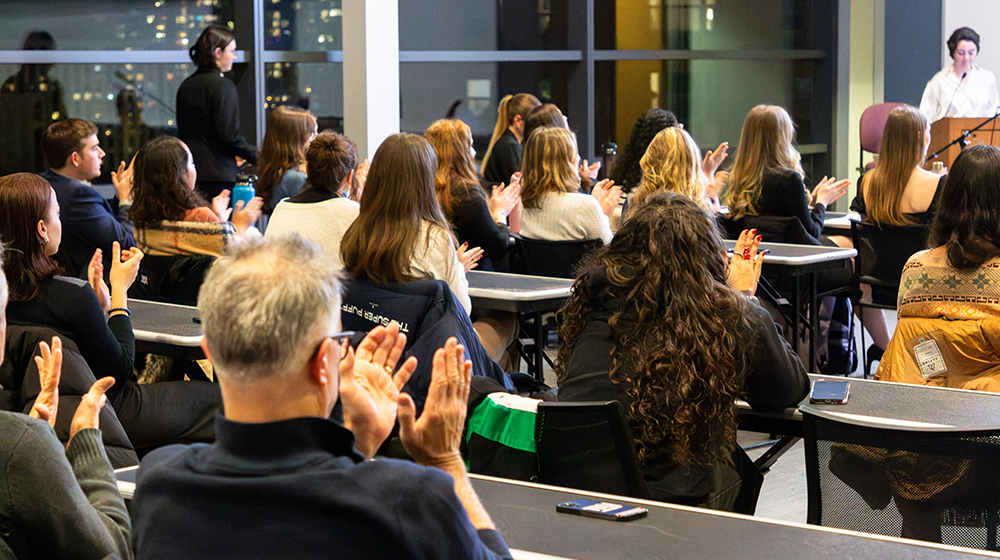 Audience seated in a classroom-style space applauds a speaker at the front of the room during an evening campus event.