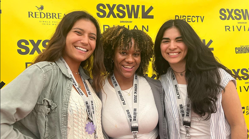 Three attendees wearing event badges pose together in front of a bright yellow SXSW backdrop featuring logos of sponsors.