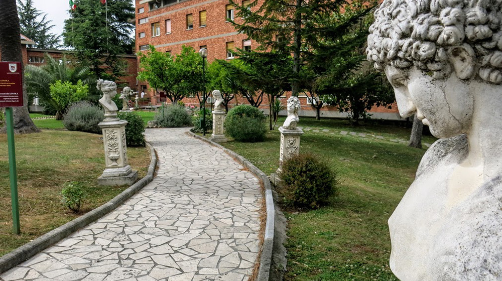 A landscaped campus walkway lined with classical bust statues leads past trees and greenery toward a brick academic building.