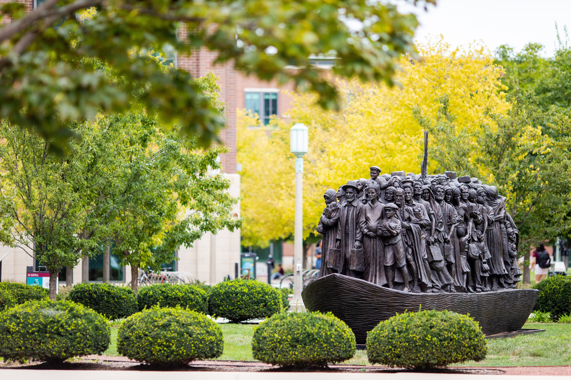 Bronze sculpture of migrants standing together in a boat on Loyola’s campus lawn, surrounded by trees and greenery.