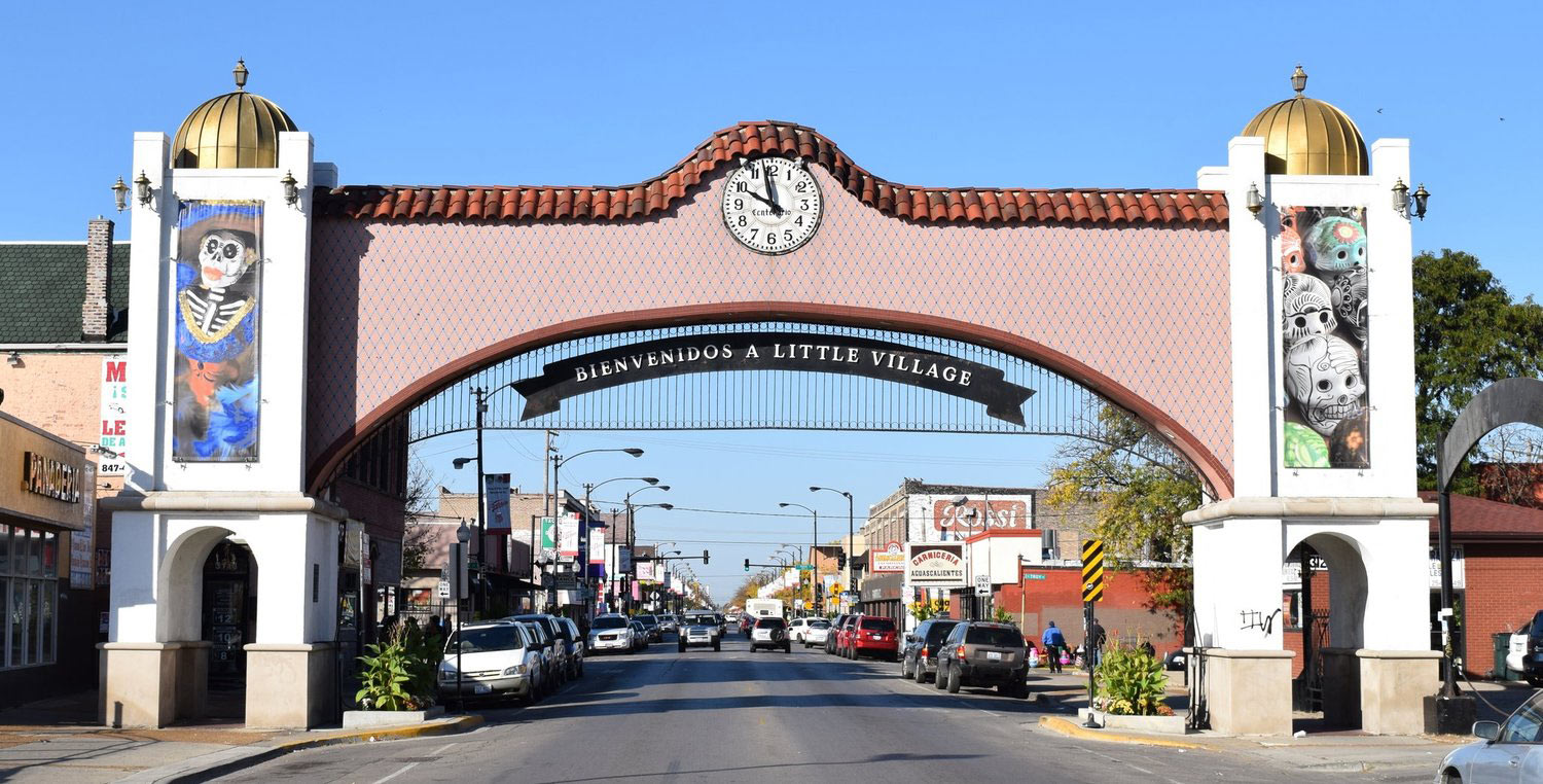 “Bienvenidos a Little Village” archway spans a busy Chicago street lined with shops and parked cars under a clear blue sky.