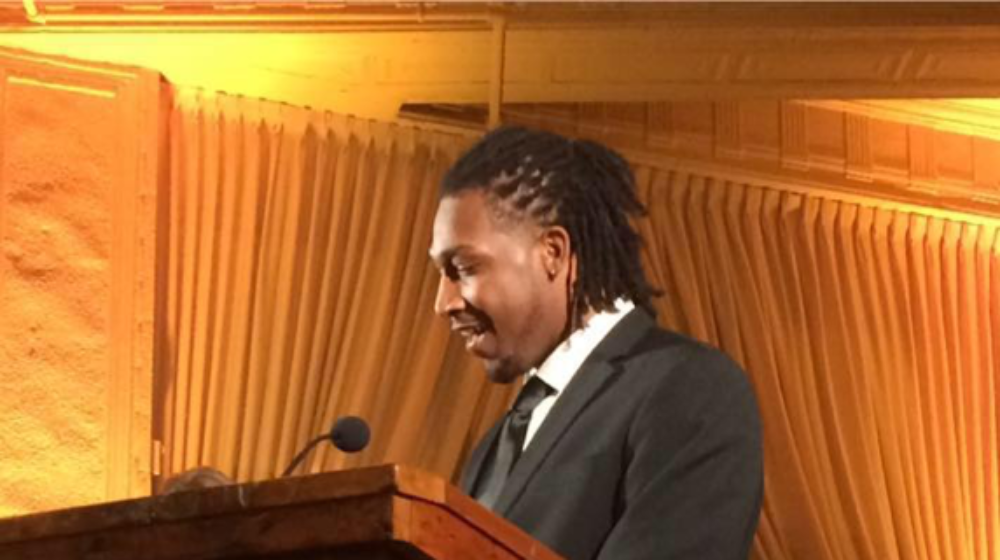 Leo Davis speaks at a podium during an indoor event, standing in front of gold curtains and a microphone.