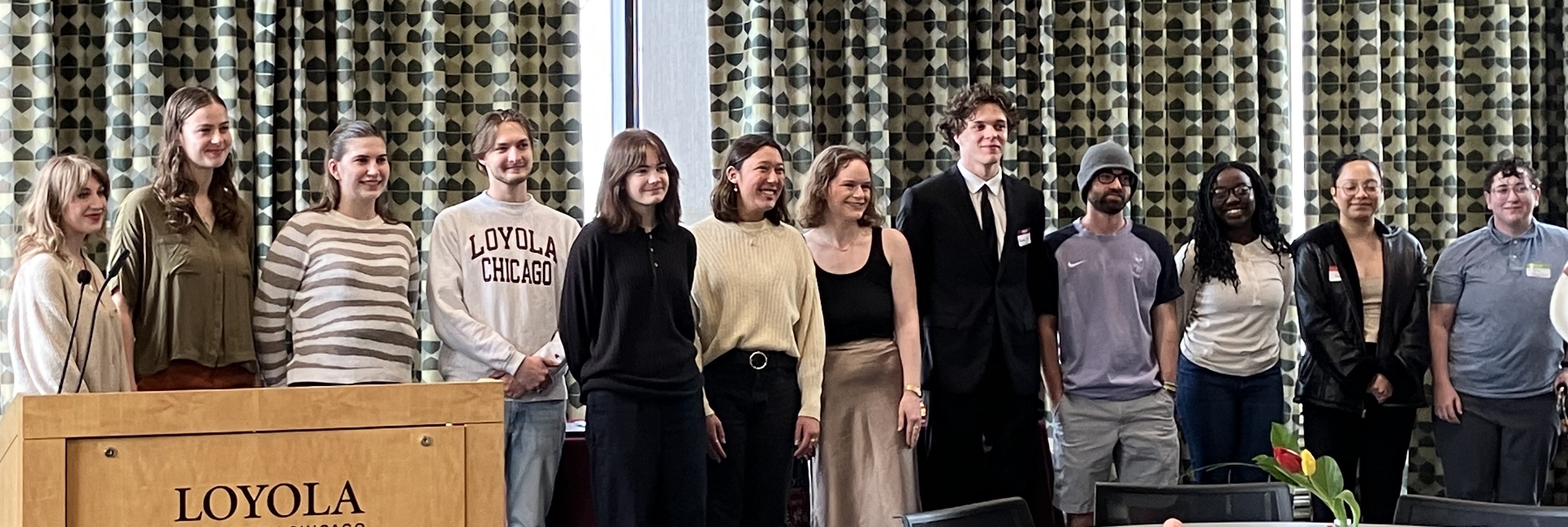 A large group of people gather in a room at large round tables and chairs for an award ceremony. Six individuals who won awards stand in front of ever