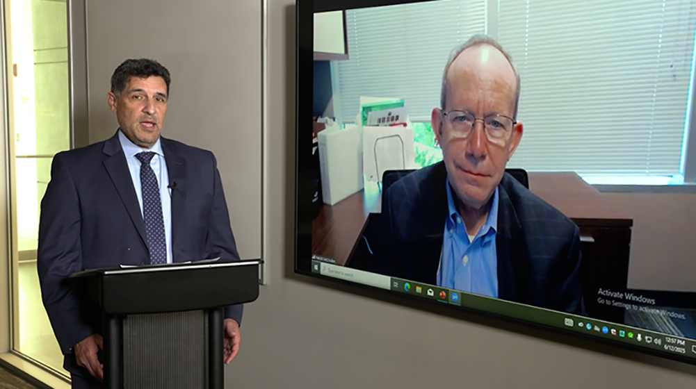Man speaking at a podium next to a screen showing a remote speaker
