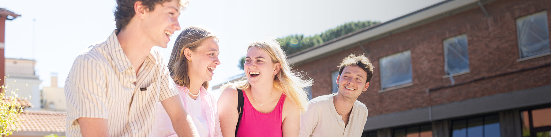 Four smiling and laughing students sit on a bench in a small plaza just outside the Rome Center.