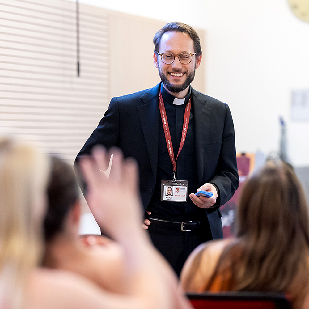 A jesuit instructor in a classroom with students smiles as he interacts with the group of students.