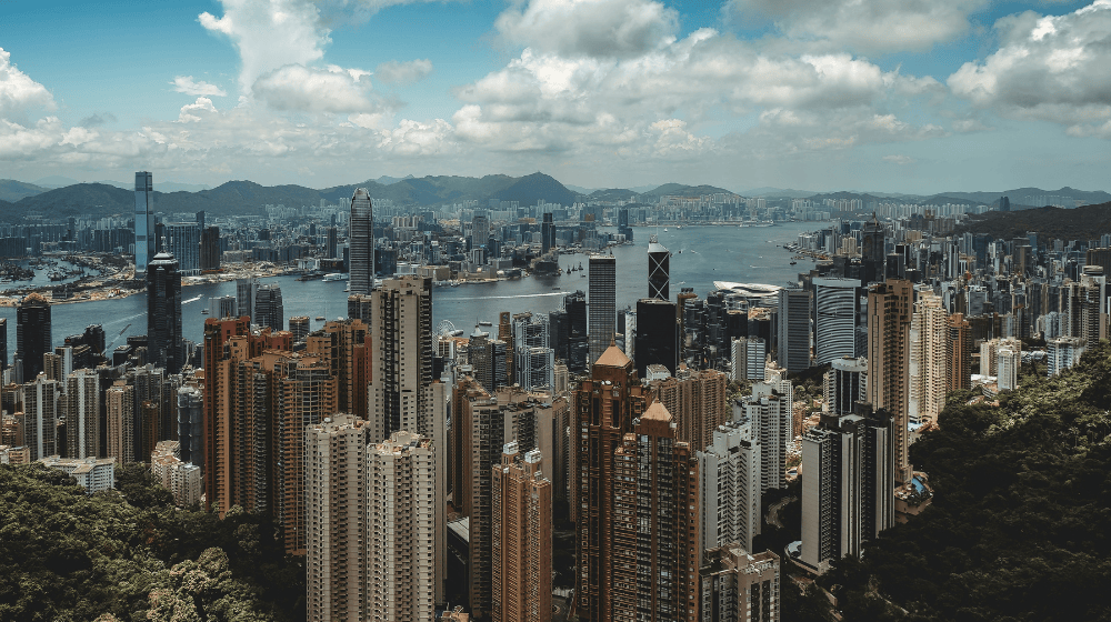 A panoramic daytime view of Hong Kong’s skyline from above, with tall skyscrapers surrounding Victoria Harbour and mountains in the background under a