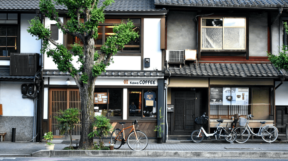A small traditional-style Japanese street with a coffee shop and bicycles parked outside, shaded by a leafy tree in front of the shop.