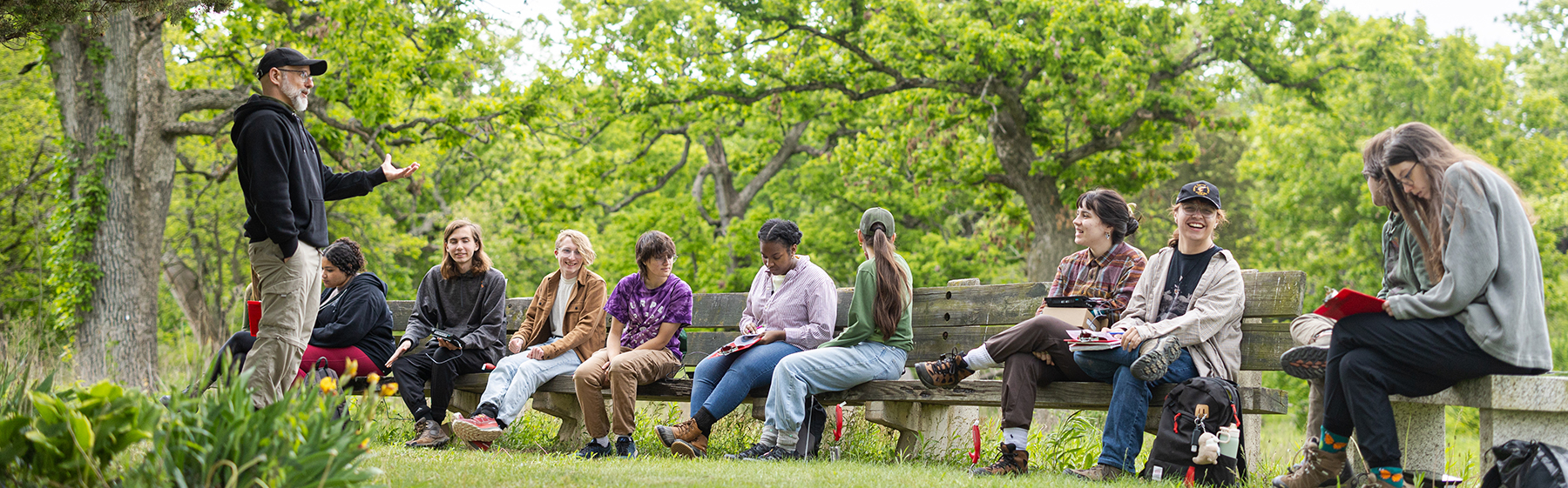 students sit outside on a bench while a professor talks