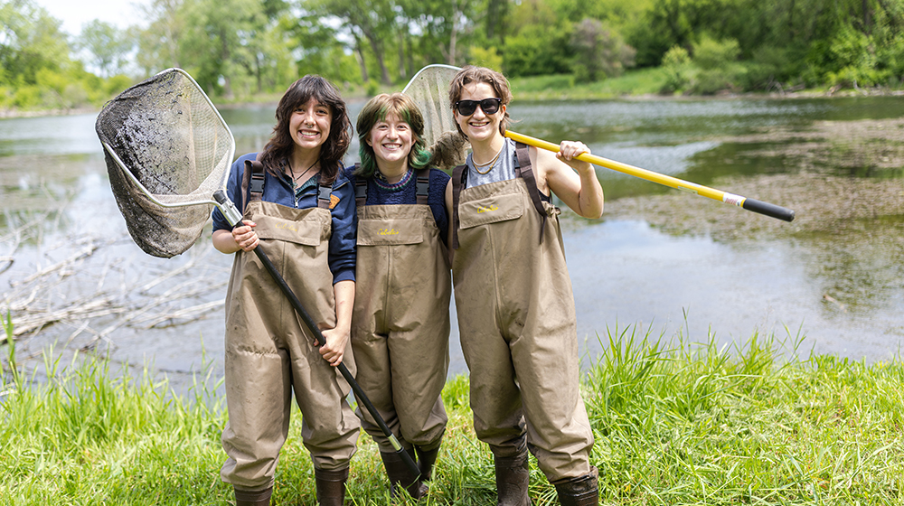 three students wearing waders smiling in front of a pond
