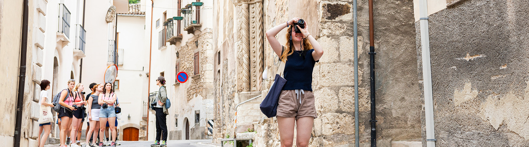 A small group of students walk together between two rows of houses as they take a study tour in Rome. One young woman with long red hair and dressed in a summer shirt and shorts, stops to take a photo. She stands several feet in front of the group on the street. She is faced away from the group.