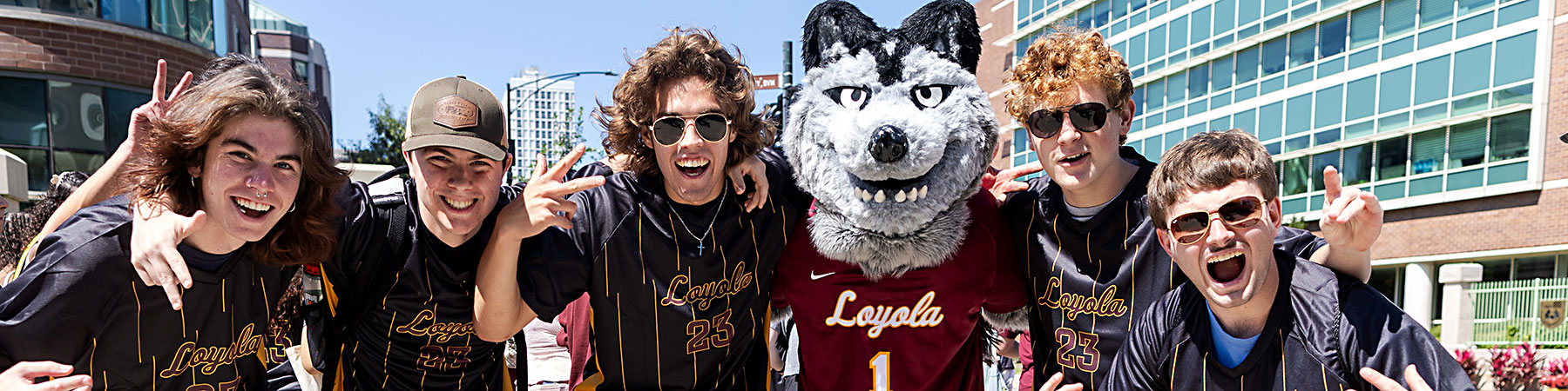 On a bright and sunny day, a group of smiling, energetic, young men surround Lu Wolf. There are several on ether side of Lu with arms up around each others shoulders.