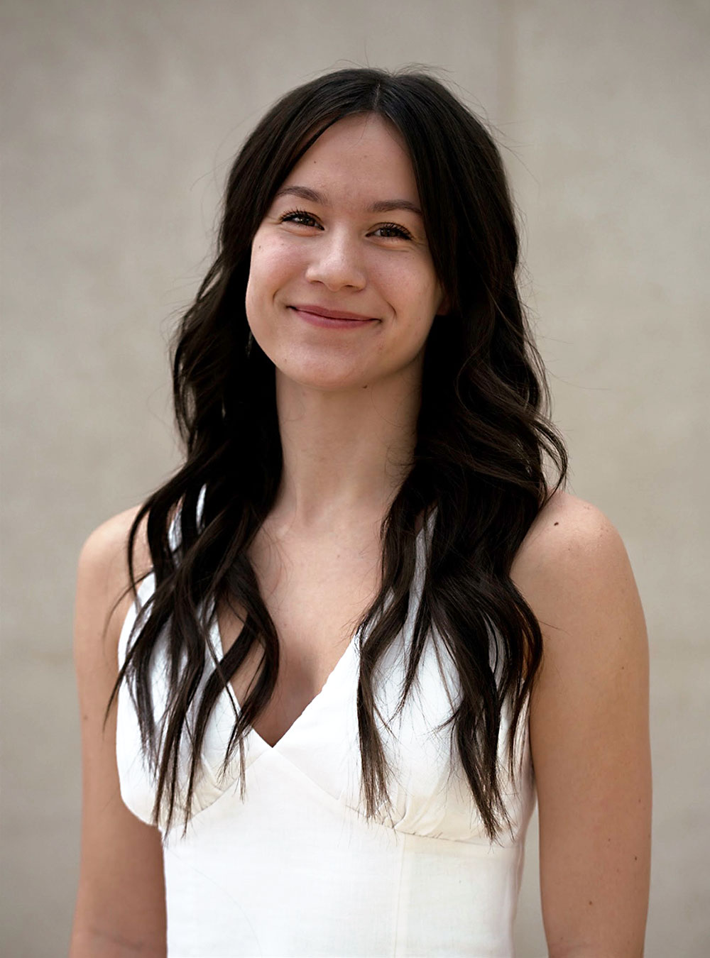 Portrait of smiling woman with long dark hair wearing a white sleeveless dress.