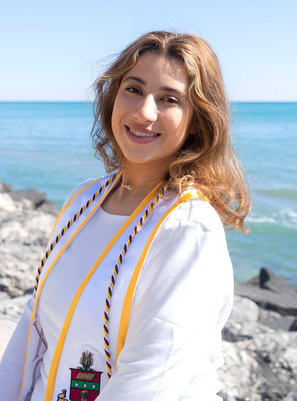 Graduate in white gown and honor cords smiling by Lake Michigan shoreline.