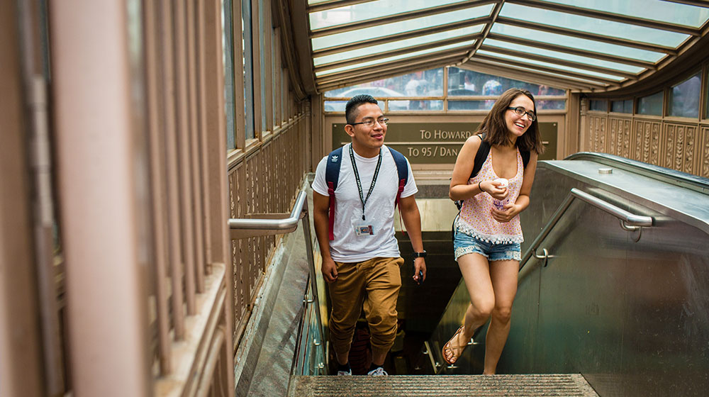 Two students with backpacks walking up subway stairs, smiling and talking.