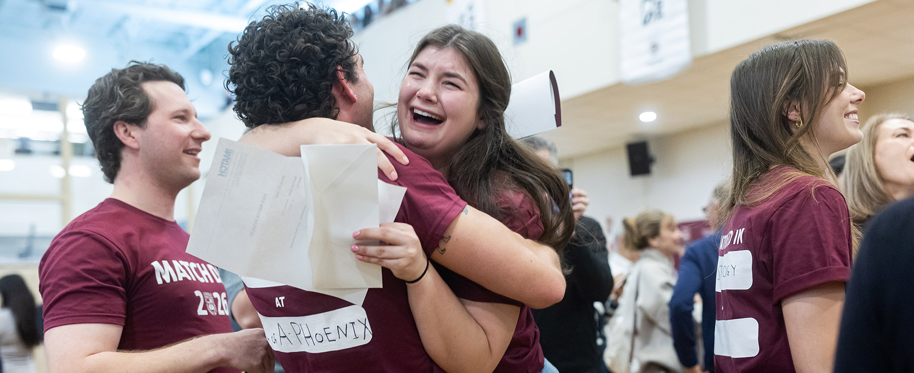 Two students wearing maroon t-shirts hugs while one cries tears of joy while surrounded by classmates, friends, and family Two students wearing maroon t-shirts hugs while one cries tears of joy while surrounded by classmates, friends, and family