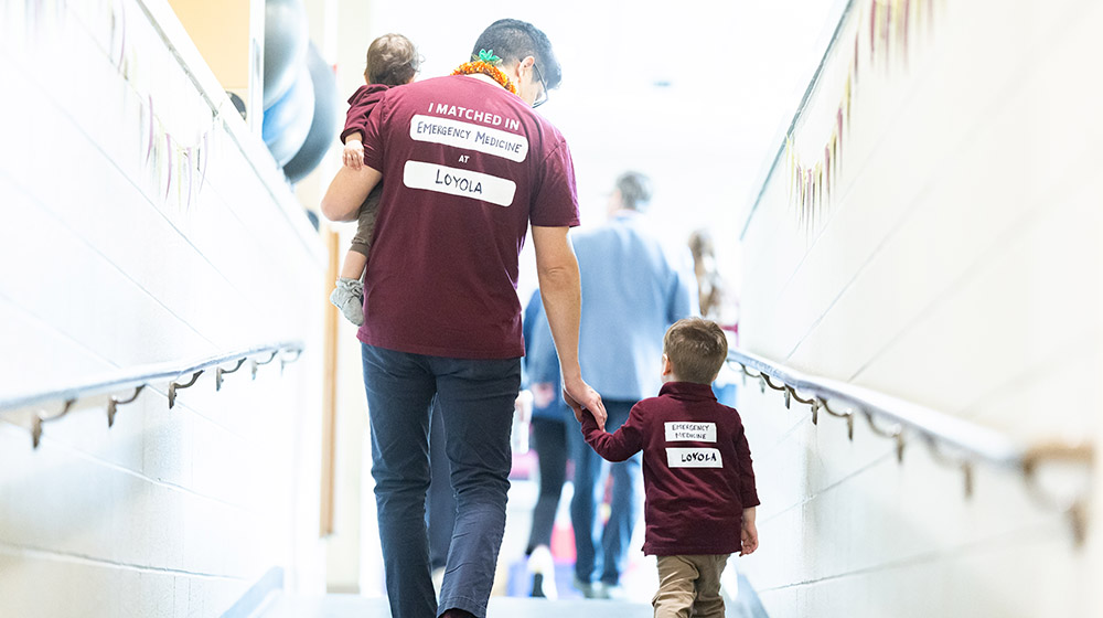 A student wearing a maroon t-shirt with writing indicating he matched in emergency medicine at Loyola walks with his two children