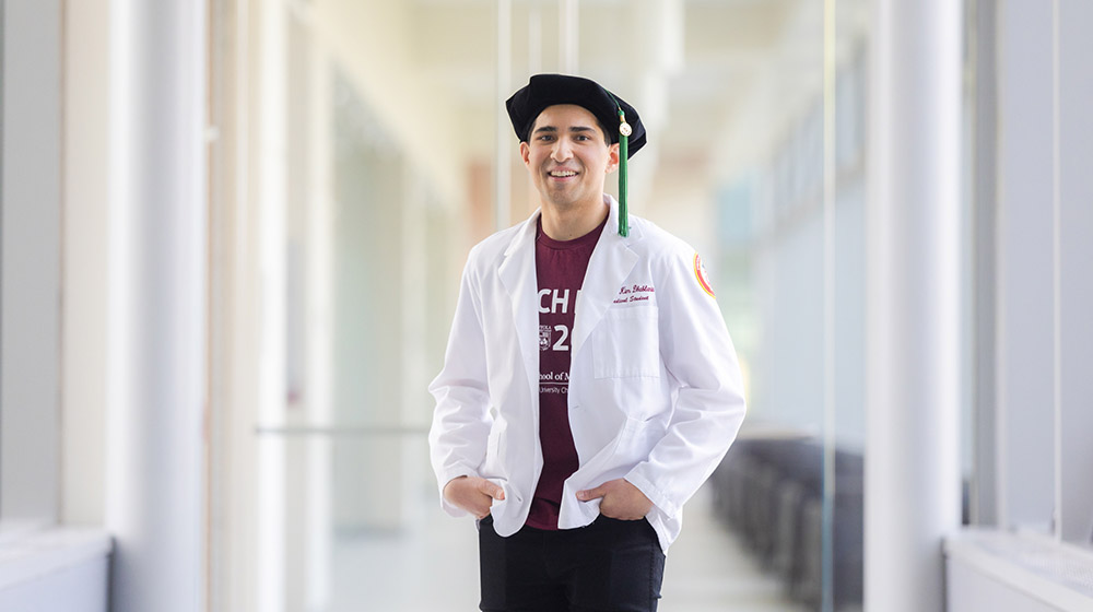 Dual MD/MPH student Nathan Dhablania standing in a hallway wearing a maroon Match Day t-shirt, his white medical coat, and his graduation cap Dual MD/MPH student Nathan Dhablania standing in a hallway wearing a maroon Match Day t-shirt, his white medical coat, and his graduation cap