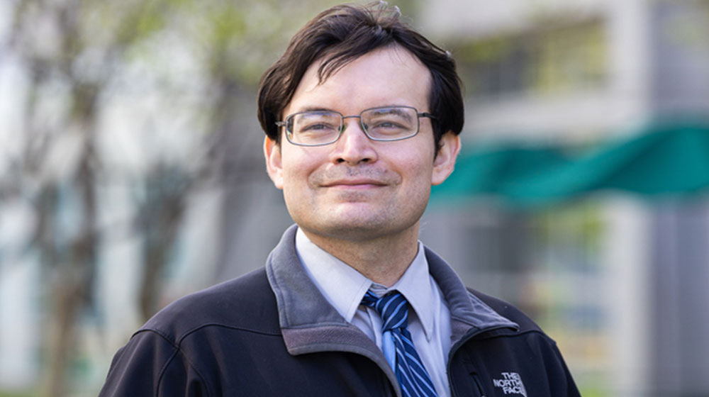 Man wearing glasses and a tie under a jacket, smiling outdoors with a blurred campus setting in the background.
